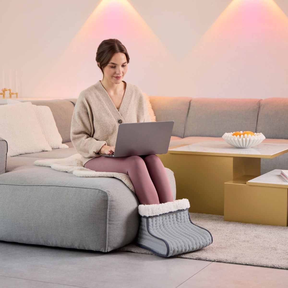 Eine Frau sitzt mit einem Laptop auf dem Sofa und trägt einen grauen elektrischen Fußwärmer in einem modernen Wohnraum.
